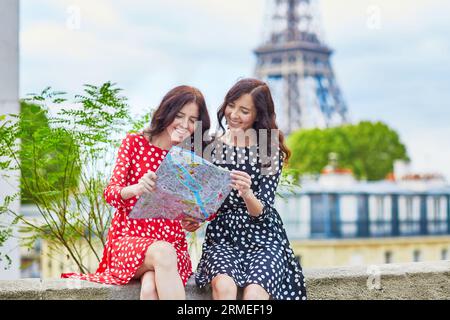 Bellissime sorelle gemelle che usano la mappa di fronte alla Torre Eiffel durante il viaggio a Parigi, in Francia. Le felici ragazze sorridenti si godono le loro vacanze in Europa Foto Stock