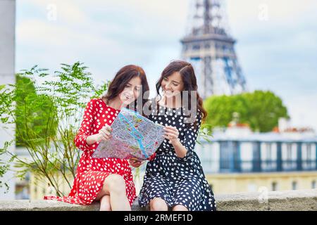 Bellissime sorelle gemelle che usano la mappa di fronte alla Torre Eiffel durante il viaggio a Parigi, in Francia. Le felici ragazze sorridenti si godono le loro vacanze in Europa Foto Stock