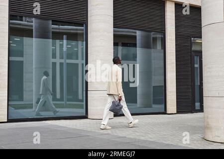 Foto lunga di un giovane uomo d'affari afroamericano in costume bianco che porta una borsa mentre cammina lungo l'architettura moderna con grandi finestre Foto Stock