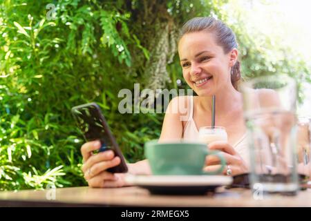 Giovane donna bruna seduta in un bar all'aperto e messaggistica online utilizzando il suo smartphone. Foto Stock