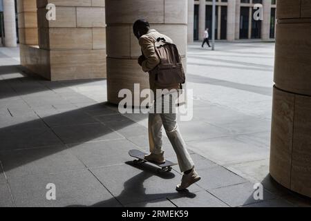 Vista posteriore di un giovane uomo d'affari con uno zaino in piedi sullo skateboard mentre cavalca per lavorare lungo enormi colonne di un edificio moderno Foto Stock