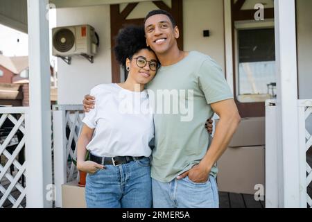 coppia afroamericana positiva che si abbraccia e posa vicino a una nuova casa sfocata sullo sfondo Foto Stock