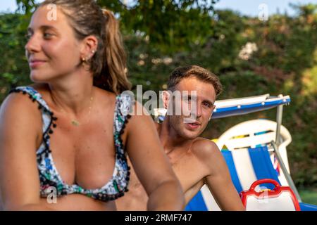 Divertente ritratto di una giovane donna incinta con marito in costume da bagno seduto su sdraio in un resort Foto Stock