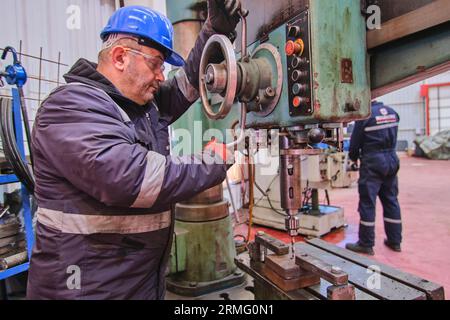 un uomo con equipaggiamento di sicurezza completo come un casco, una visiera e un giubbotto. eseguire la saldatura su un tubo di metallo arrugginito. un uomo che usa una sega elettrica portatile su un tubo metallico. Foto Stock