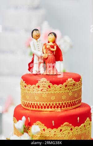 Splendida torta nuziale rossa e gialla in stile indiano con statuette per sposi e sposi in cima Foto Stock