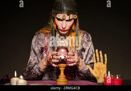 Una donna che indossa accessori seduti al tavolo con candele accese e oggetti che raccontano la fortuna mentre consulti la sfera di cristallo durante la D. Foto Stock