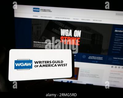 Persona che possiede uno smartphone con il logo del sindacato Writers Guild of America West (WGAW) sullo schermo di fronte al sito Web. Concentrarsi sul display del telefono. Foto Stock