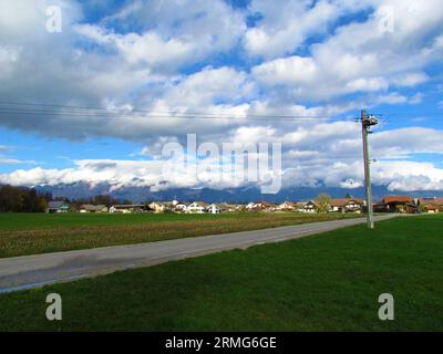 Vista del villaggio vicino a Kranj a Sorsko polje nella regione di Gorenjska in Slovenia con le alpi Kamnik-Savinja nella parte posteriore coperte da nuvole e le case in f Foto Stock