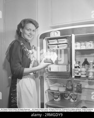 In cucina anni '1950 All'interno di una cucina e di una giovane donna in piedi al frigiratore Leonard con la porta aperta, che mostra le lattine e le bottiglie di cibo diverso in piedi sugli scaffali. Nel frigorifero è integrato un armadietto di congelamento separato, una caratteristica abbastanza nuova in questo momento. Indossa un grembiule di plastica. È Haide Göransson, 1928-2008, modella e attrice. Svezia 1950 Kristoffersson rif AU23-2 Foto Stock