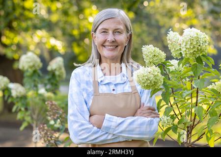 Ritratto di una donna in pensione giardiniera, anziana casalinga dai capelli grigi che sorride e guarda la macchina fotografica, con le braccia incrociate, che tende al giardino di casa in campagna, con ortensie di fiori. Foto Stock
