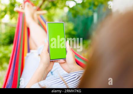 Donna sdraiata su un'amaca in giardino, tiene lo smartphone in mano con un cromakey Foto Stock