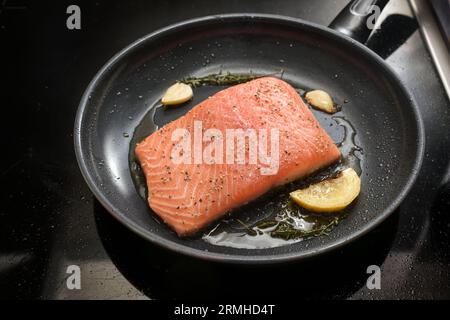 Il filetto di salmone fresco viene fritto sulla pelle con limone, erbe aromatiche e aglio in una padella nera sul piano cottura, spazio di copia, messa a fuoco selezionata, profondità stretta o Foto Stock