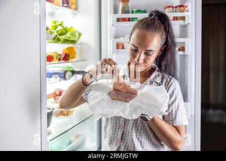 Donna molto affamata in pigiama che si godeva il prosciutto di notte vicino al frigo. Foto Stock