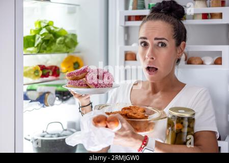 Il venerdì sera, una donna affamata di pigiama sceglie spuntini, bistecche, ciambelle e una lattina di sottaceti dal frigorifero. Foto Stock