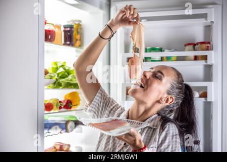 Donna molto affamata in pigiama che si godeva il prosciutto di notte vicino al frigo. Foto Stock