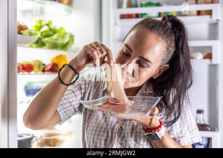 Donna molto affamata in pigiama che si godeva il prosciutto di notte vicino al frigo. Foto Stock