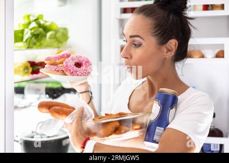 Il venerdì sera, una donna molto affamata in pigiama sceglie spuntini, snack, ciambelle e una lattina di birra dal frigorifero. Foto Stock