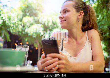 Vista dall'alto, giovane bruna seduta estiva in una caffetteria all'aperto con il telefono cellulare in mano. Foto Stock