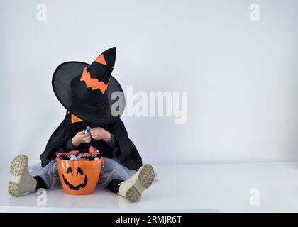 Bambina vestita da strega in cerca di caramelle nel cestino di halloween, sfondo bianco Foto Stock