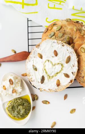 Colazione con carboidrati molto gustosa e salutare. Bagel appena sfornato con formaggio cremoso, semi di girasole e cuore dipinto condito con salsa al pesto Foto Stock