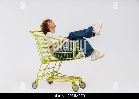 Allegra donna di mezza età seduta nel carrello della spesa isolata su sfondo bianco Foto Stock