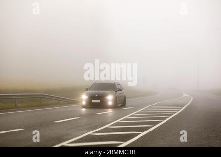 Auto BMW nera con fari luminosi che guidano su strada attraverso la fitta nebbia mattutina. Salo, Finlandia. 25 agosto 2023. Foto Stock