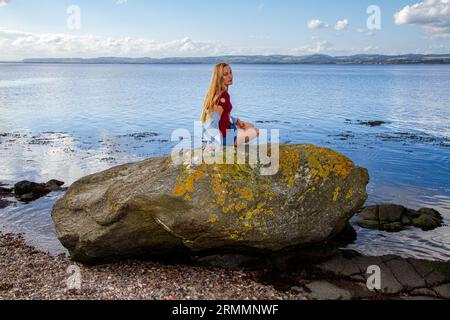 Una bella donna bionda, Rhianna Martin, siede sulle rocce della spiaggia di Wormit nella contea di Fife, in Scozia, godendo del clima piacevole, Regno Unito Foto Stock