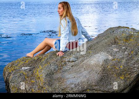 Una bella donna bionda, Rhianna Martin, siede sulle rocce della spiaggia di Wormit nella contea di Fife, in Scozia, godendo del clima piacevole, Regno Unito Foto Stock