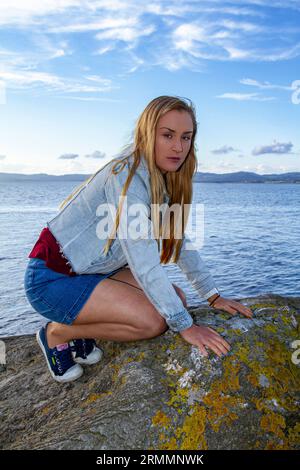 Una bella donna bionda, Rhianna Martin, siede sulle rocce della spiaggia di Wormit nella contea di Fife, in Scozia, godendo del clima piacevole, Regno Unito Foto Stock