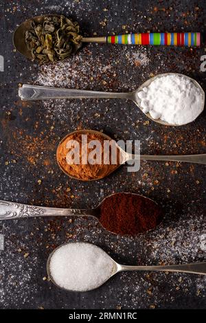 Cucchiai con tè verde, cacao, zucchero a velo e caffè macinato su fondo nero cosparso di zucchero di canna e bianco, caffè e cacao in polvere. Foto Stock