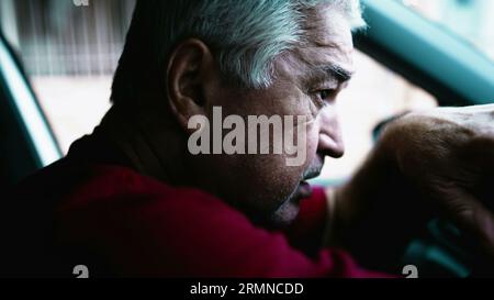 Uomo anziano che soffre di disperazione emotiva parcheggiato in auto, persona anziana che si appoggia al volante del veicolo in una quieta disperazione e disconnessione sociale Foto Stock