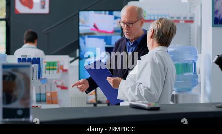 Acquirente anziano che guarda la pubblicità di prodotti farmaceutici sullo schermo digitale in farmacia, si avvicina al farmacista con l'intenzione di acquistare farmaci pubblicizzati. Esperto assiste il cliente senior con informazioni Foto Stock