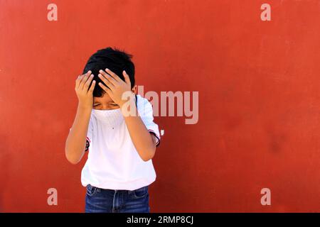 Felice e penoso Latino 8 anni ragazzo che indossa camicia bianca uniforme scolastica e maschera viso per la protezione nella pandemia di Covid-19 al ritorno a scuola Foto Stock