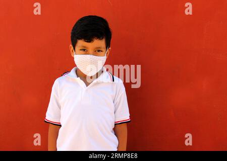 Felice e penoso Latino 8 anni ragazzo che indossa camicia bianca uniforme scolastica e maschera viso per la protezione nella pandemia di Covid-19 al ritorno a scuola Foto Stock