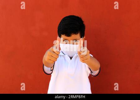 Felice e penoso Latino 8 anni ragazzo che indossa camicia bianca uniforme scolastica e maschera viso per la protezione nella pandemia di Covid-19 al ritorno a scuola Foto Stock