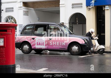 Un tradizionale taxi nero londinese con un annuncio pubblicitario, accanto a un pilota di scooter con consegna di cibo, con un ciclista dietro e una cabina rossa. Londra, Regno Unito Foto Stock