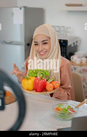 Un ritratto di una bella blogger di cibo musulmana asiatica o influencer di stile di vita sta mostrando gli ingredienti del suo pasto sano alla fotocamera mentre Foto Stock