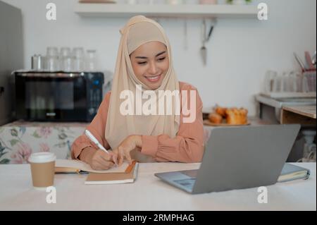 Una bella studentessa asiatica musulmana sta facendo i compiti o studiando per una lezione online a un tavolo in cucina, scrivendo qualcosa Foto Stock