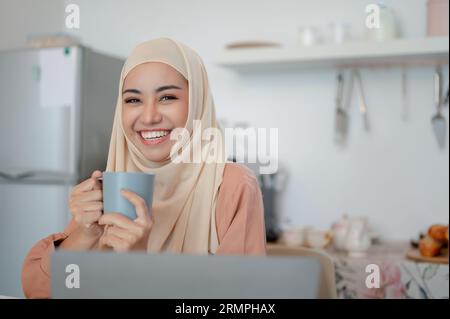 Una bella e sorridente donna musulmana asiatica che indossa l'hijab siede a un tavolo in cucina con una tazza di caffè in mano e un portatile sul tavolo, lavora Foto Stock