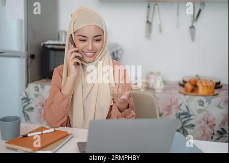 Una bella donna musulmana asiatica con un hijab o un velo parla al telefono mentre lavora al suo computer portatile a un tavolo in cucina. lavorare da casa, Foto Stock