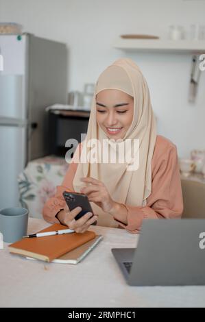 Un'attraente donna musulmana asiatica che indossa un hijab sta usando il suo smartphone e risponde ai messaggi a un tavolo in cucina. concetto di vita domestica Foto Stock