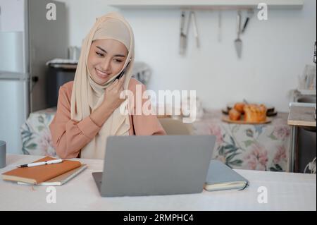 Una bella donna musulmana asiatica con un hijab o un velo parla al telefono mentre lavora al suo computer portatile a un tavolo in cucina. lavorare da casa, Foto Stock