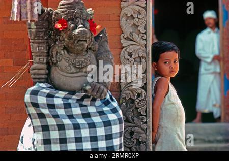 Statua guardiana del tempio di Bedogol decorata con fiori di ibisco, una timida bambina e sacerdote sullo sfondo davanti a un tempio a Gianyar, Bali, Indonesia Foto Stock