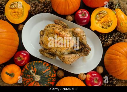 Delizioso pollo arrosto su un tavolo per la cena del giorno del Ringraziamento, circondato da zucche, mele, noci e coni. Composizione stagionale autunnale. Foto Stock