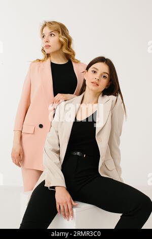 Modella le donne vestite con blazer e pantaloni pastello alla moda con sneakers eleganti che si posa su un cubo su uno sfondo bianco nello studio. Foto Stock