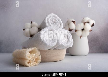 Stack of rolled white towels with cotton flower and loofah sponge on gray background Stock Photo