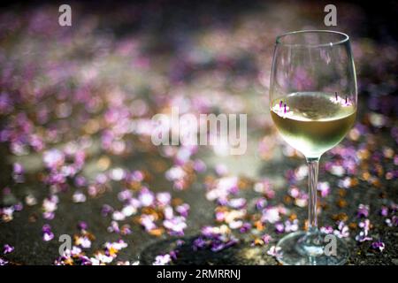 Un bicchiere di vino bianco, con fiori lilla caduti sullo sfondo Foto Stock