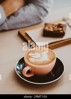 Cappuccino o latte con schiuma spugnosa in una tazza di caffè in ceramica e torta brownie sullo sfondo del tavolo. Caffè e bar, cappuccino, tazza di caffè nel caffè. Foto Stock