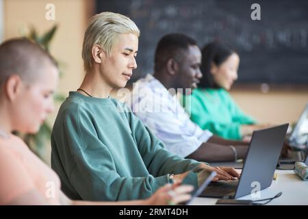Focus on young Asian male student or programmer using laptop while sitting among intercultural classmates or coworkers at college Foto Stock