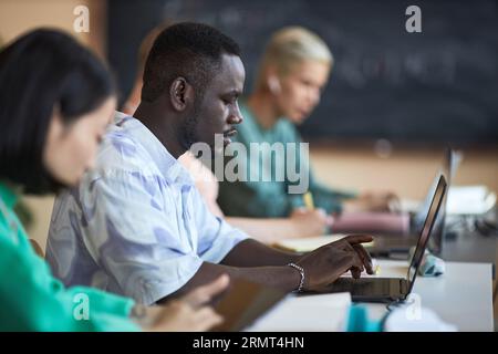 Vista laterale di un giovane studente afroamericano delle scuole superiori che utilizza un computer portatile mentre è seduto tra compagni di classe interculturali e decodifica dei dati Foto Stock